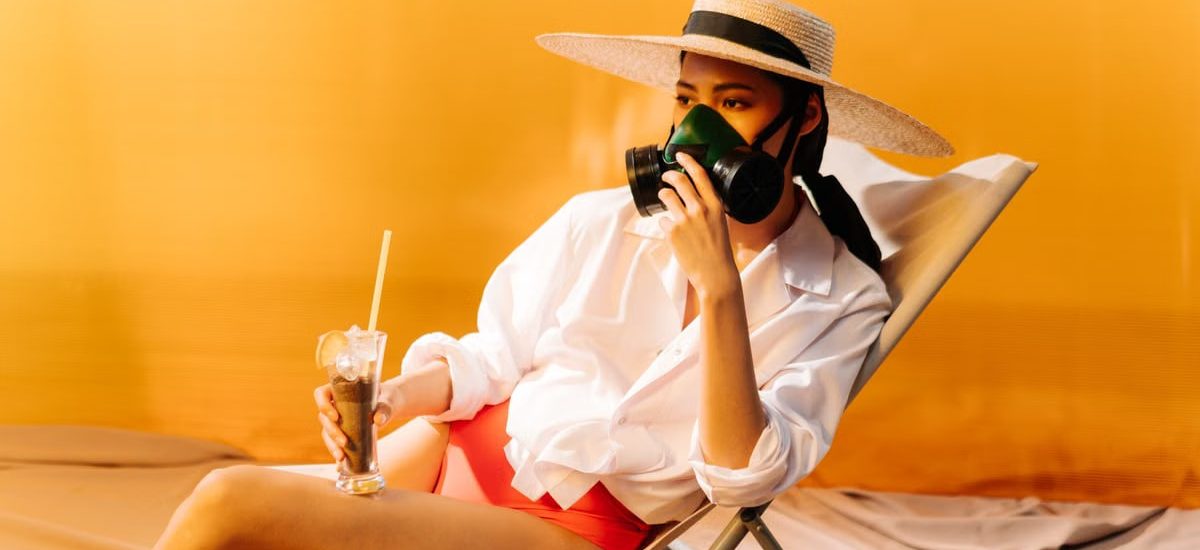 Woman wearing a gas mask and sun hat while sitting on a chair with a drink, symbolizing climate change.