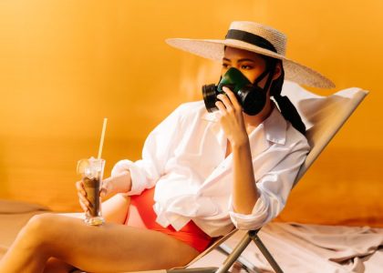 Woman wearing a gas mask and sun hat while sitting on a chair with a drink, symbolizing climate change.